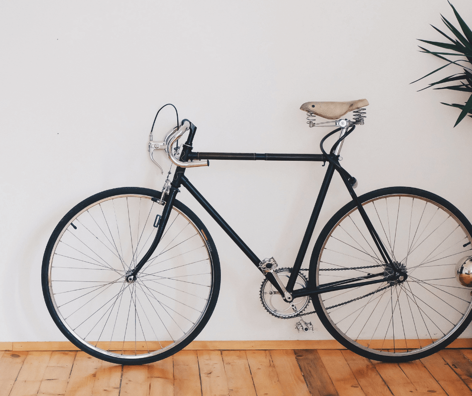 A black, fixed gear bicycle with a beige seat rests against a white wall inside a wooden floored room in a house.