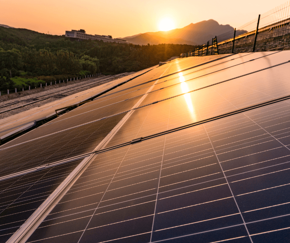 A long line of solar panels on land near trees and mountains at dusk.