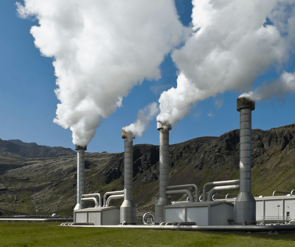 Steam rises out of four metal pillars on a geothermal power station built next to a rocky hill in the countryside.
