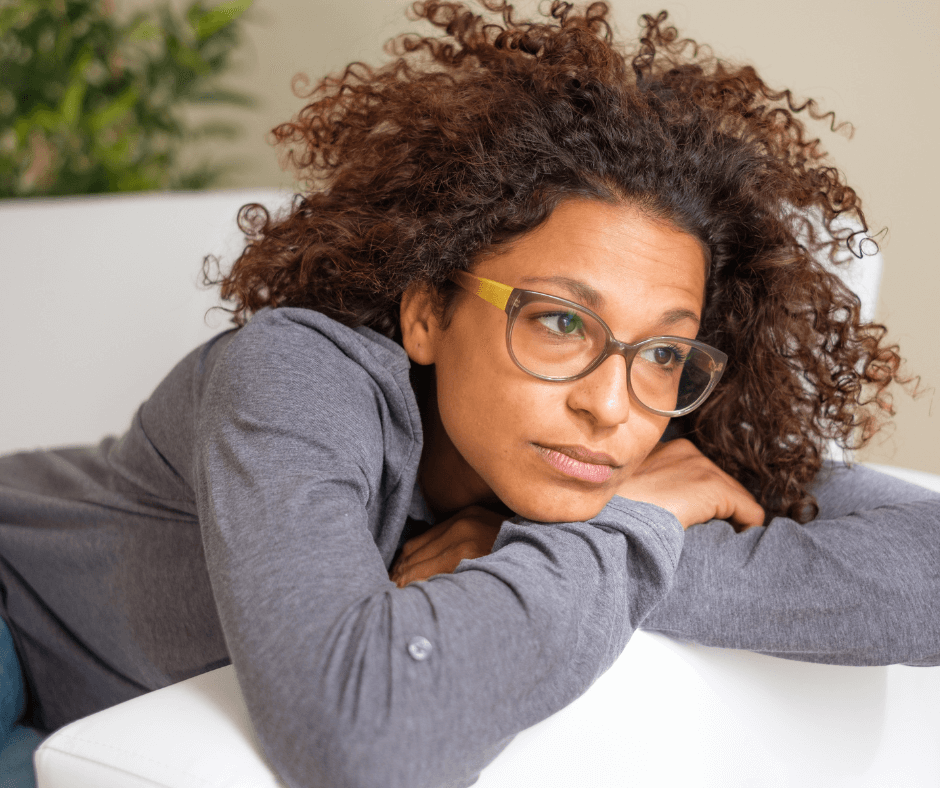 A Black woman with a worried expression on her face leans on the arm of her couch with her chin resting on her crossed arms.