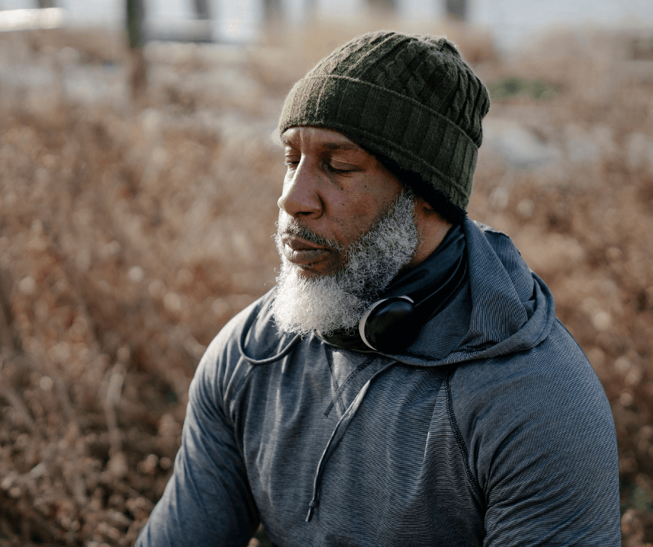 A Black man with a grey beard wearing a green beanie hat and grey hoodie sits outside with his eyes closed in quiet, relaxing contemplation.