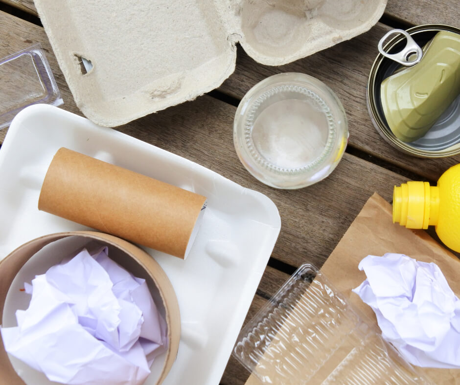 A collection of recyclable household items (tins, plastic bottles, paper, and cardboard) laid out on a wooden floor.