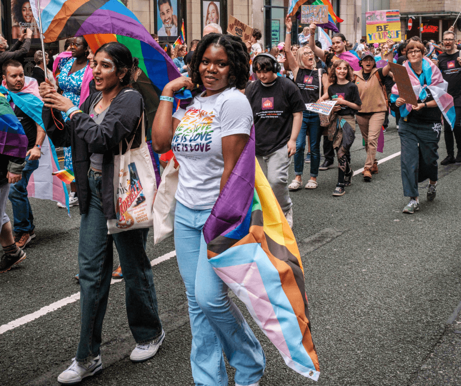 A group of people participating in a pride parade march down a city street. They carry large rainbow and transgender pride flags. The person in the foreground, wearing a white shirt with 'LOVE IS LOVE' written in rainbow colors, smiles while holding a large, progress pride flag over their shoulder.