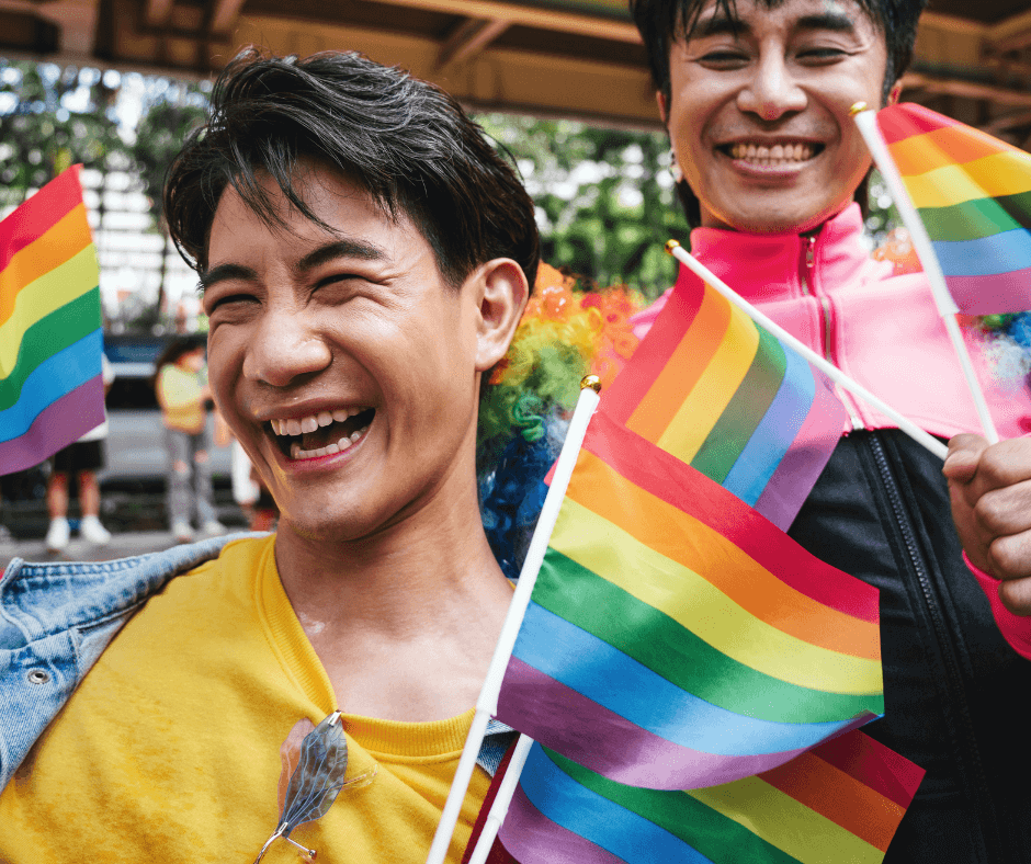 Two young Asian men hold small Rainbow Pride flags during an LGBTQ+ parade.