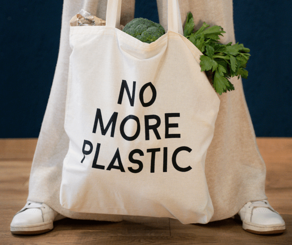 A large beige tote bag filled with fresh produce has the words “No More Plastic” written on it in big, bold black letters.