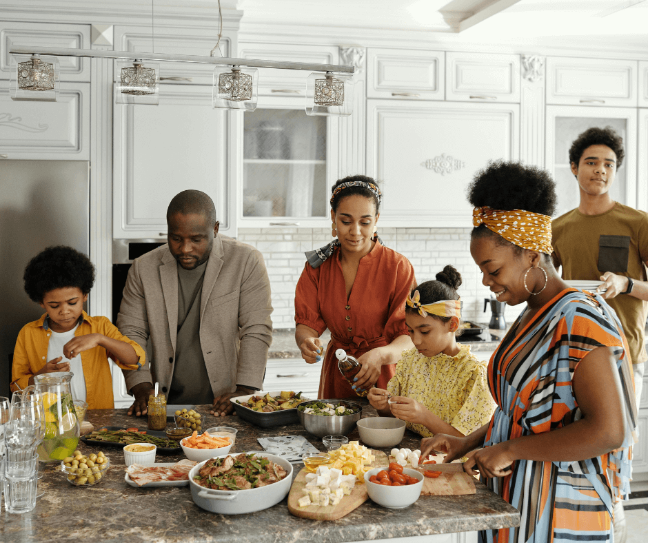 Six members of a happy Black family, varying in age from young child to adult, prepare dinner together in a white and white marble kitchen.