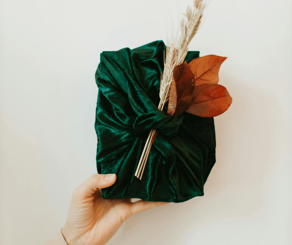 A hand holds up a holiday present sustainably wrapped in a dark green material with some dried, natural foliage tucked into the knot at the front.