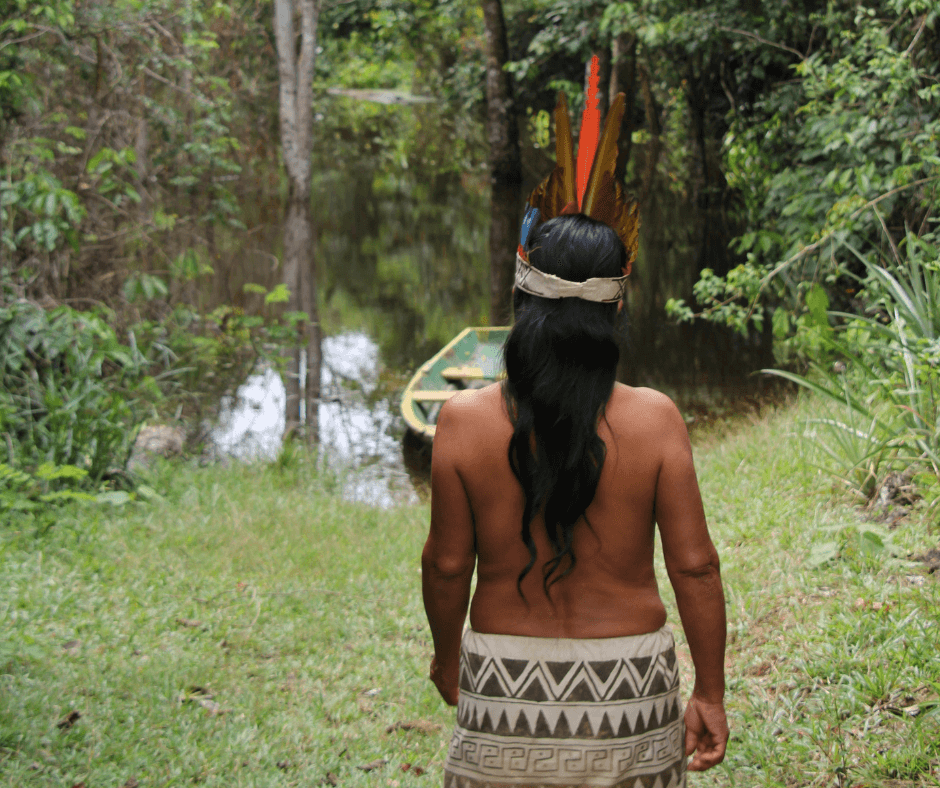 An Indigenous woman from Iquitos, Peru, walks towards a wooden canoe on small body of water in the middle of a forest; she is wearing a traditional headdress and skirt.