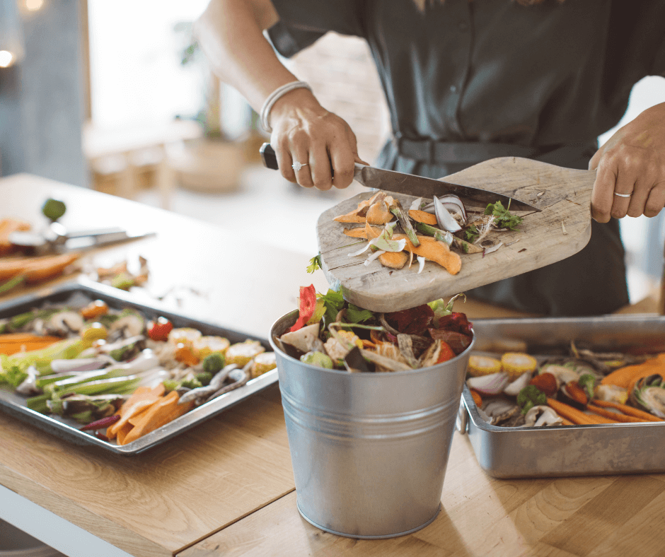 A person scraping vegetable scraps into a container to reuse them later to reduce food waste, with trays of fresh vegetables on a wooden counter in the background.