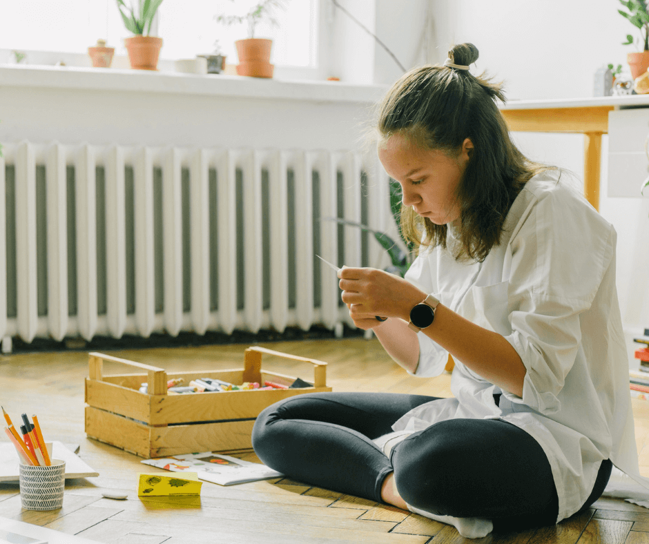 A young teenage girl sits crossed legged on a wooden floor doing an art project with various art supplies around her, including paper and pencils.