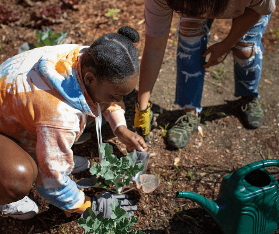 In a sunny outdoor garden, a Black teenage girl crouches down to plant something in the soil. Nearby is a green plastic watering can.
