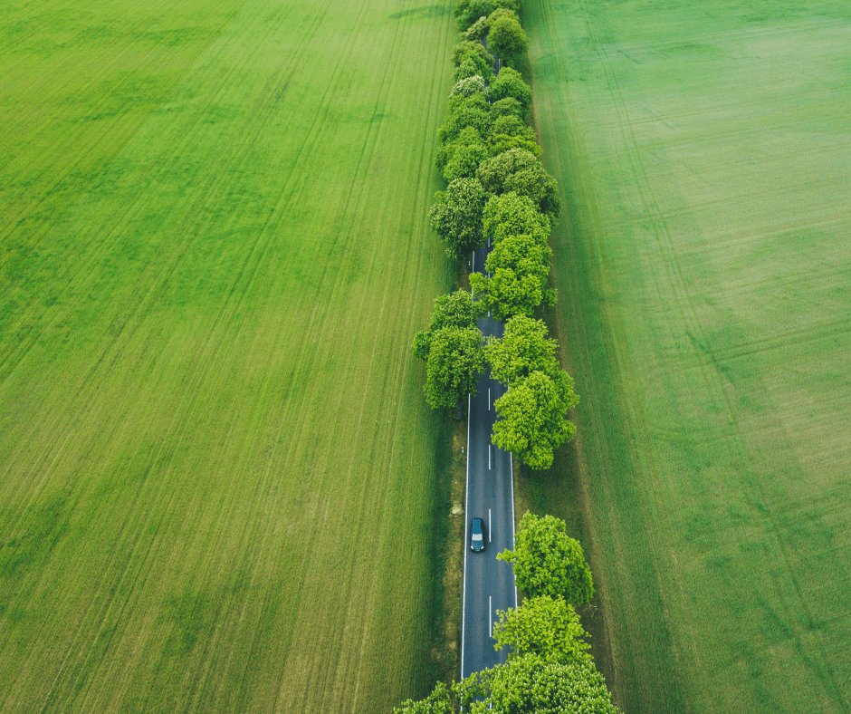 A two lane-road, shot from above, that runs through lush, green fields and trees has one electric car being driven on it.