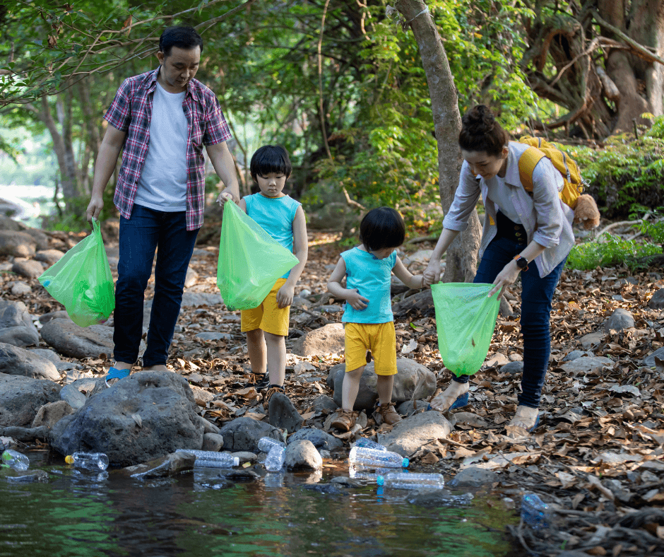An Asian family, consisting of two adults and two small children, stands next to a small river picking up plastic waste and putting it into green bags.