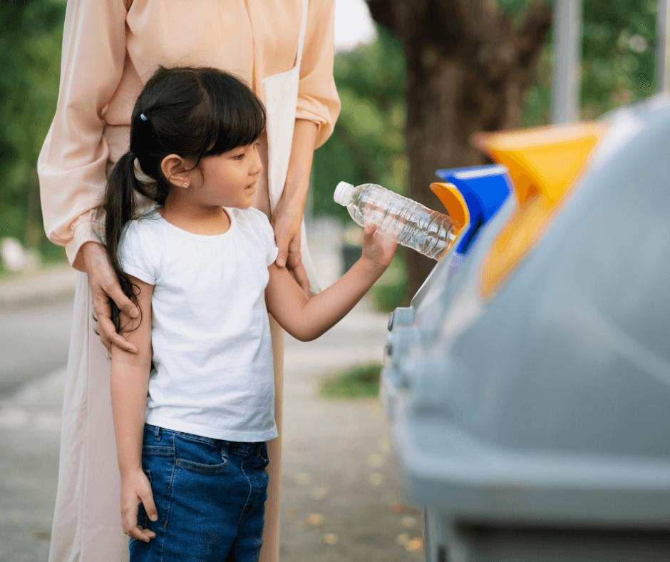 A young Asian girl about four years old places a plastic bottle in a recycling bin with the help of an adult female standing behind her. The girl has her straight, black hair in a ponytail and wears a plain white t-shirt and blue jeans.