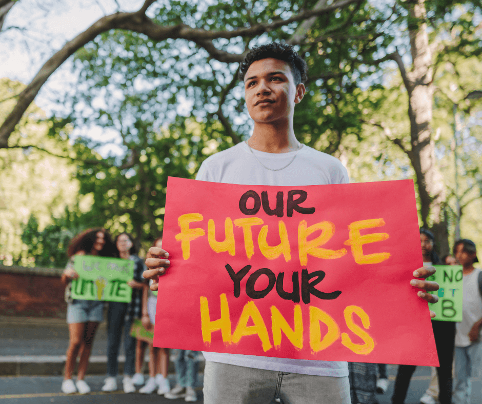 An older teenage boy with short, thick dark brown hair stands outside near a road holding a climate change protest sign made of red cardboard that reads, "Our Future, Our Hands".