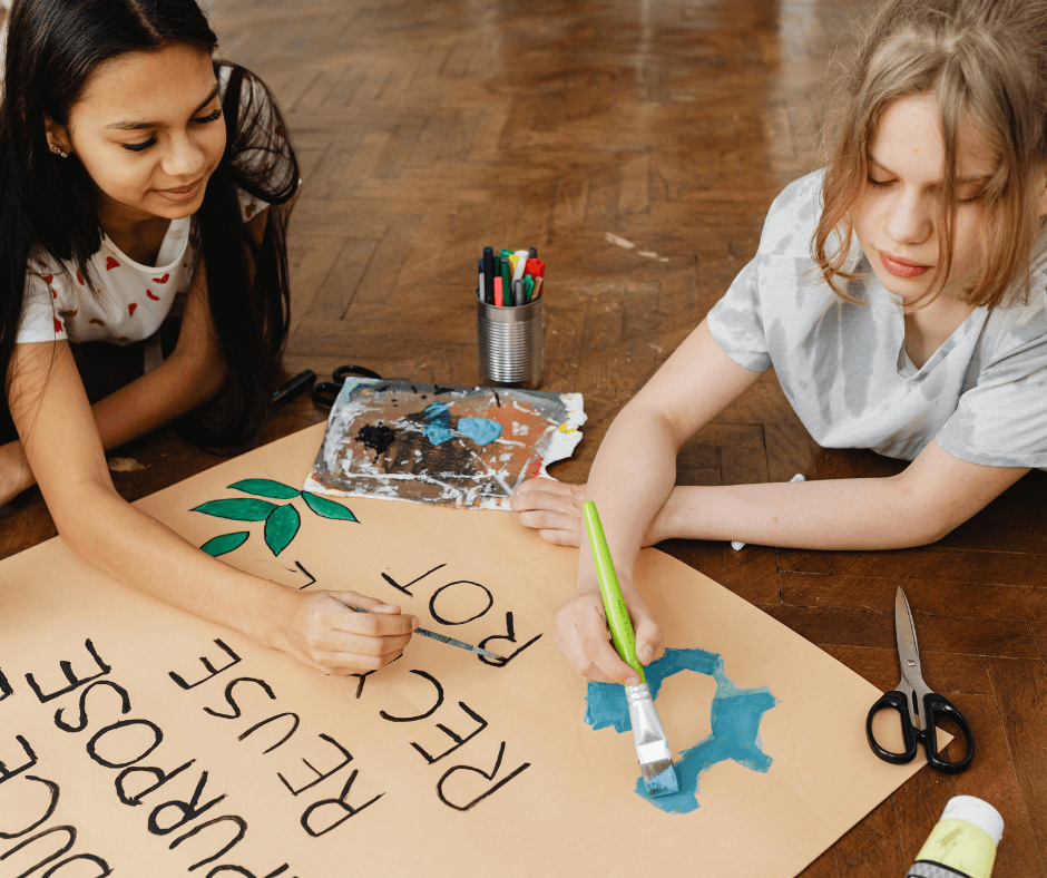 Two young teenage girls, possibly aged about 14 years old, lay propped up on a wooden floor making a poster to remind people to reuse, repurpose, and recycle.