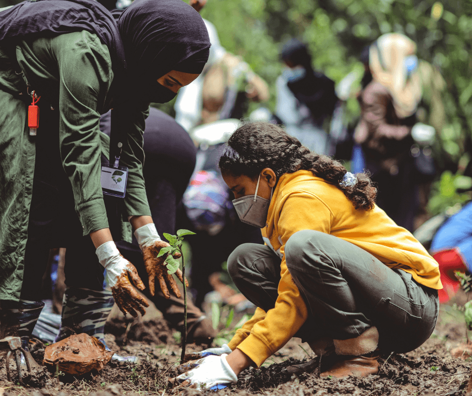 A woman wearing a black hijab, face mask, and gloves, together with a girl also wearing a face mask and gloves, plant a young tree in a wooded area.